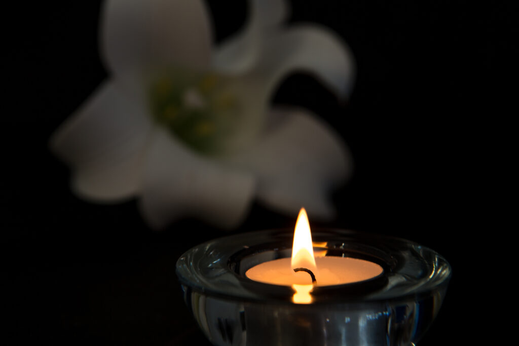 Tea light candle lighting in glass holder with white lily in background symbolising loss